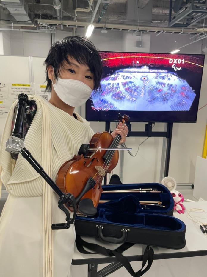 A woman, who uses a prosthetic arm, lifts a violin and poses in front of a screen showing the Opening Ceremony of the Tokyo 2020 Paralympic Games.