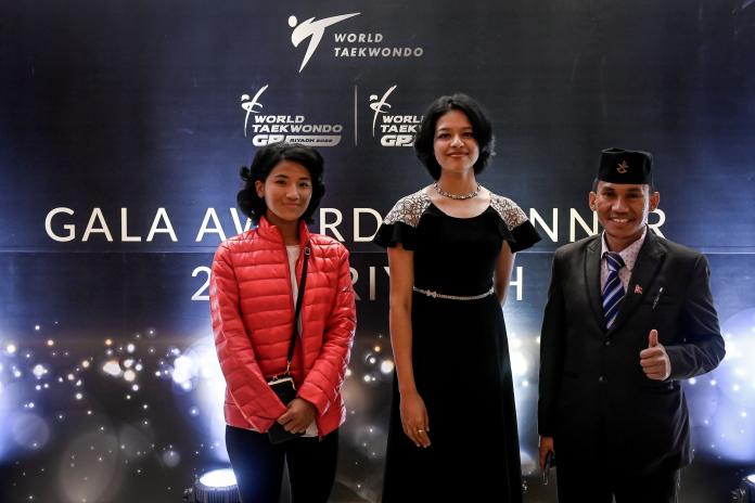 Two female athletes, one in an evening dress and the other in a sports jacket, and a male coach pose next to the official Gala Awards Dinner backdrop.