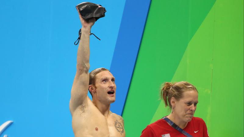 a male Para swimmer holds up his swimming cap in victory