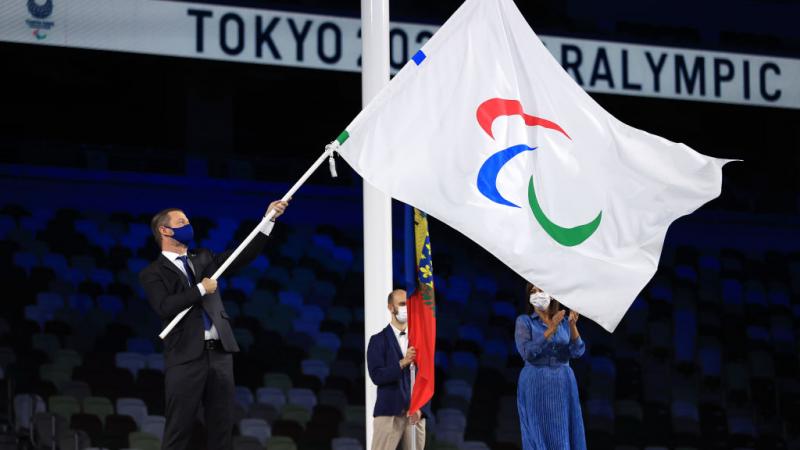 A man waves a flag with the Agitos logo on a stage at a stadium as a man and woman look on.