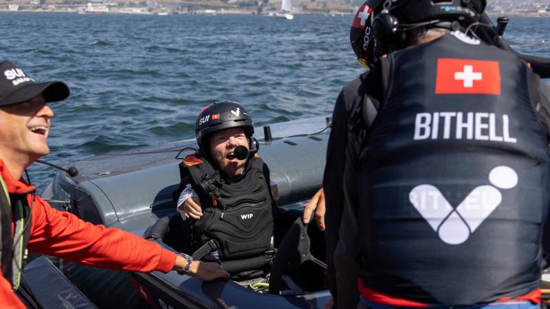 A man in a lifejacket smiles widely inside a sailboat.
