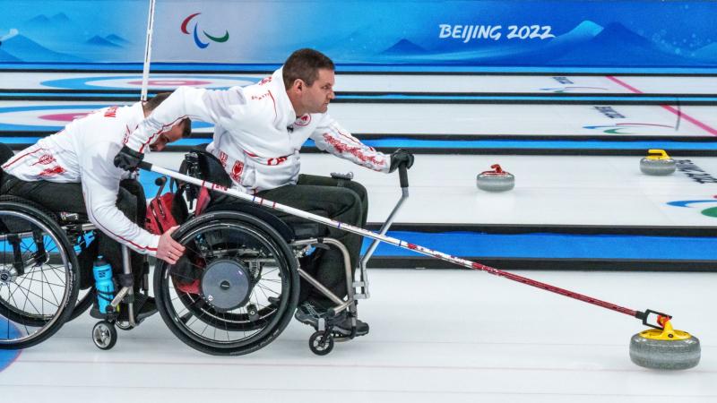 A male athlete slides a curling stone using a stick on the ice at Beijing 2022. He is on a wheelchair and another male athlete is holding onto the tires.