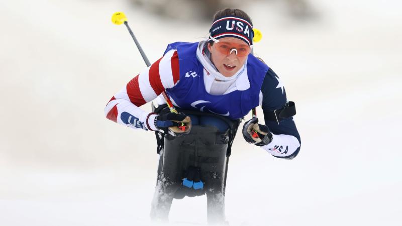 A female athlete competes in a sit-ski. She is wearing a head band that says USA and uniform in red, white and blue.
