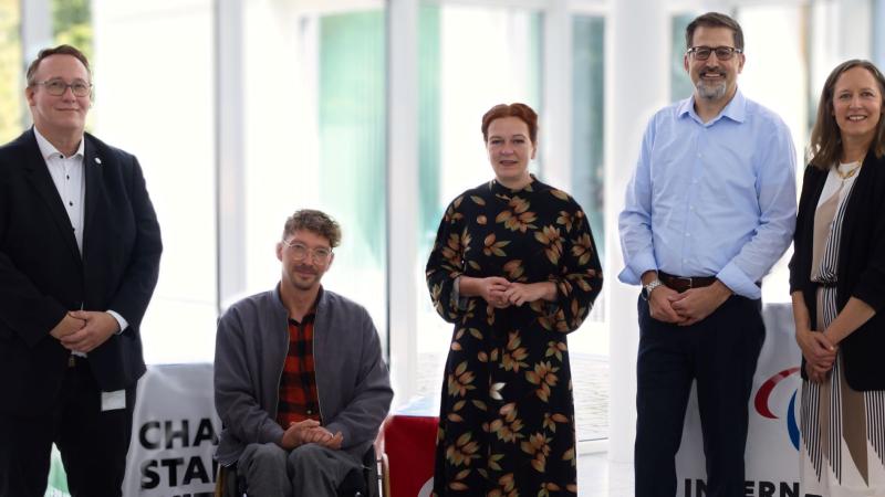 Five people pose for a photo in front of glass windows at the IPC headquarters in Bonn, Germany