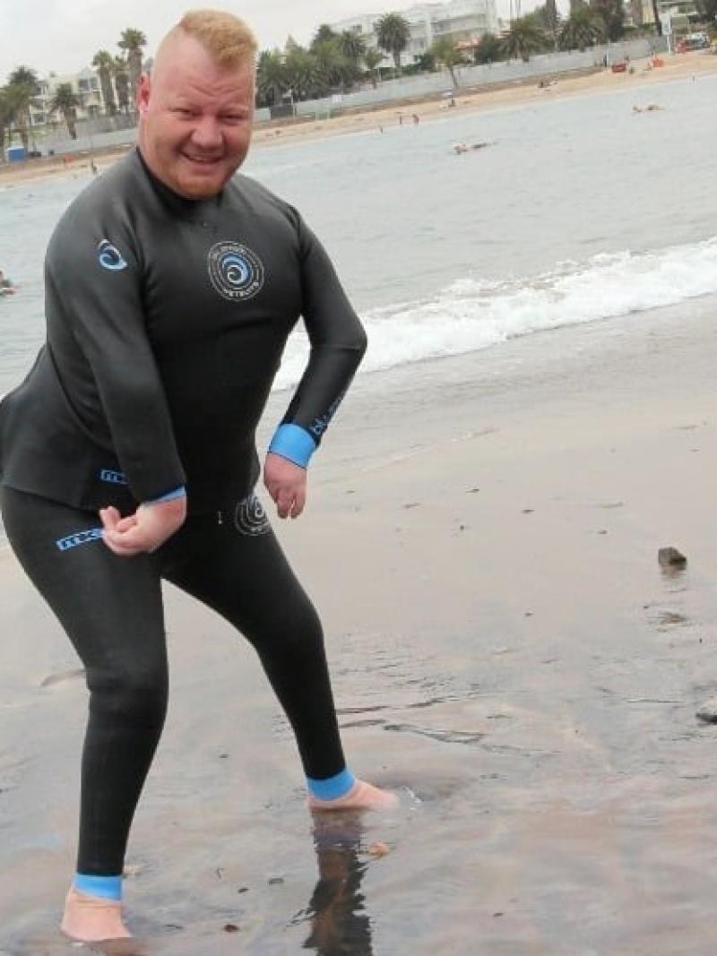 A male in a wet suit stands on the ocean shore, his feet in the water.