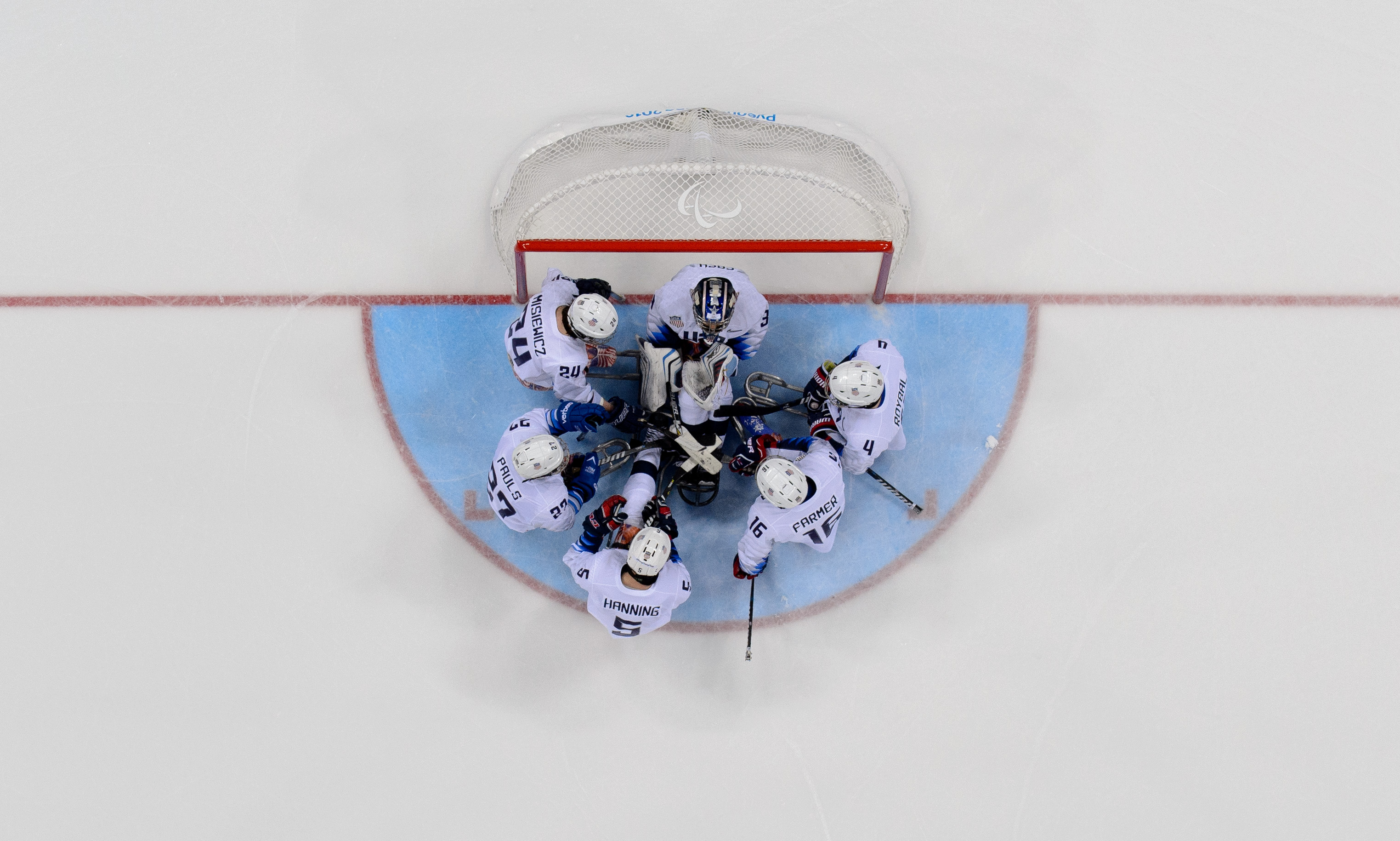 A group of six Para ice hockey players  huddle together in front of the goal