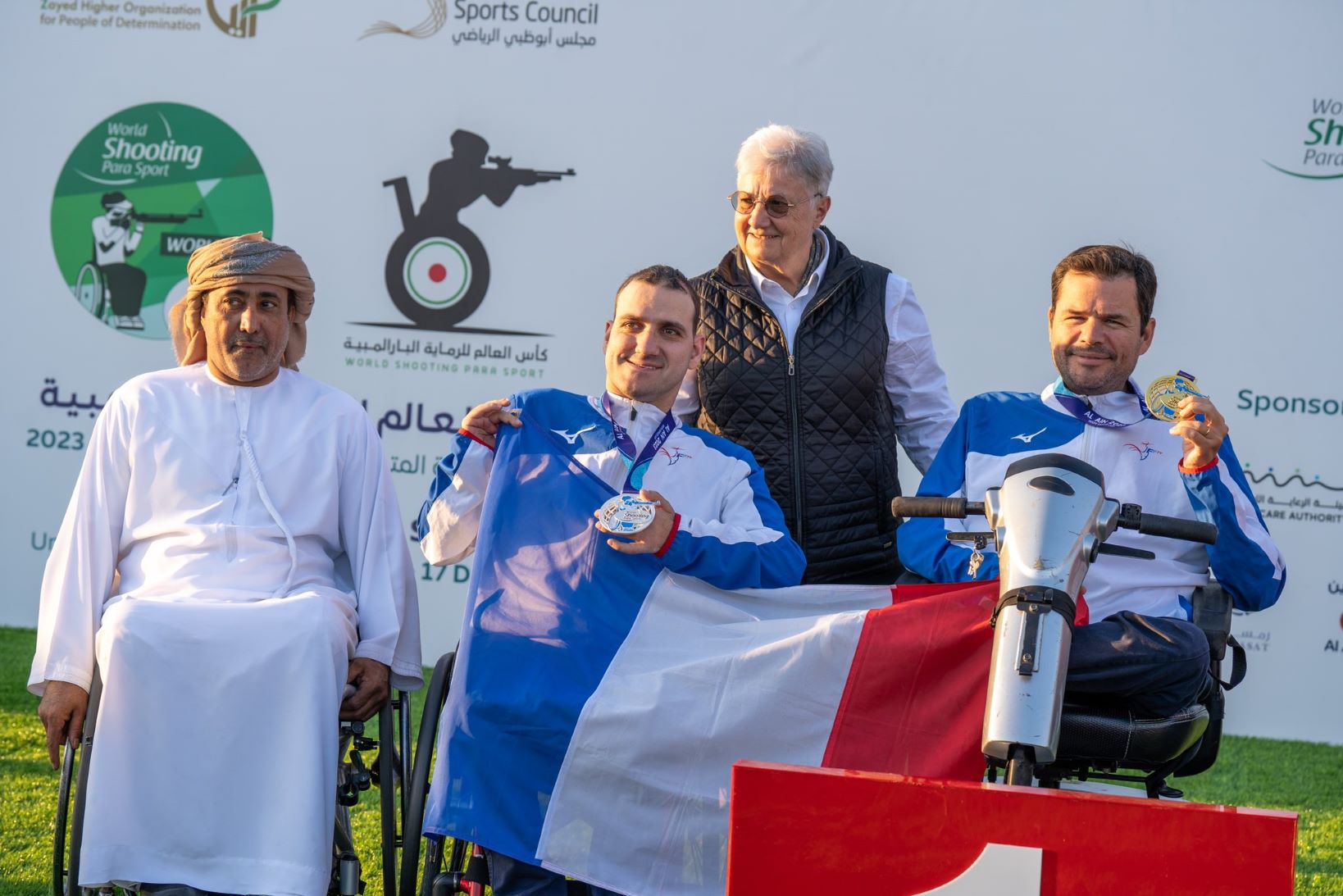 Two men in wheelchairs holding a French flag next to a man in a wheelchair and a woman standing behind them