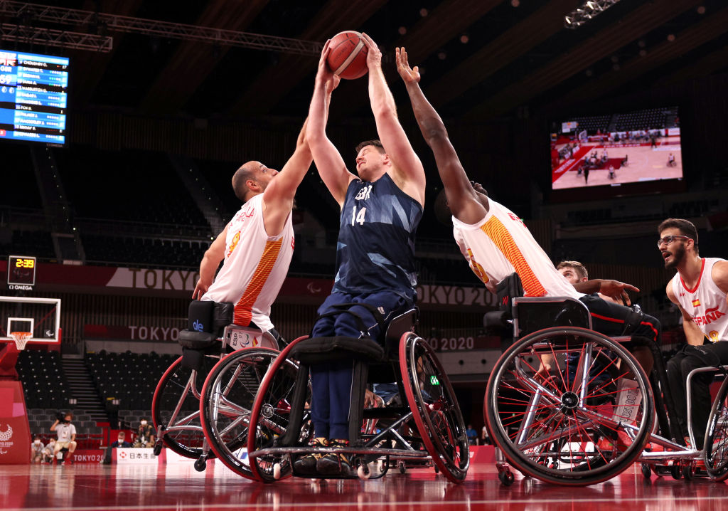 A male wheelchair basketball player wearing a blue jersey tries to shoot, while two players wearing a white jersey are reaching out to block him.