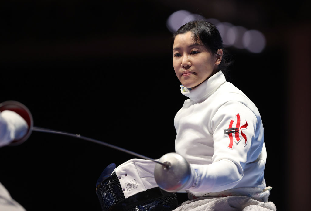 A female wheelchair fencer strikes a pose. She is holding her sword with her left and her mask with her right