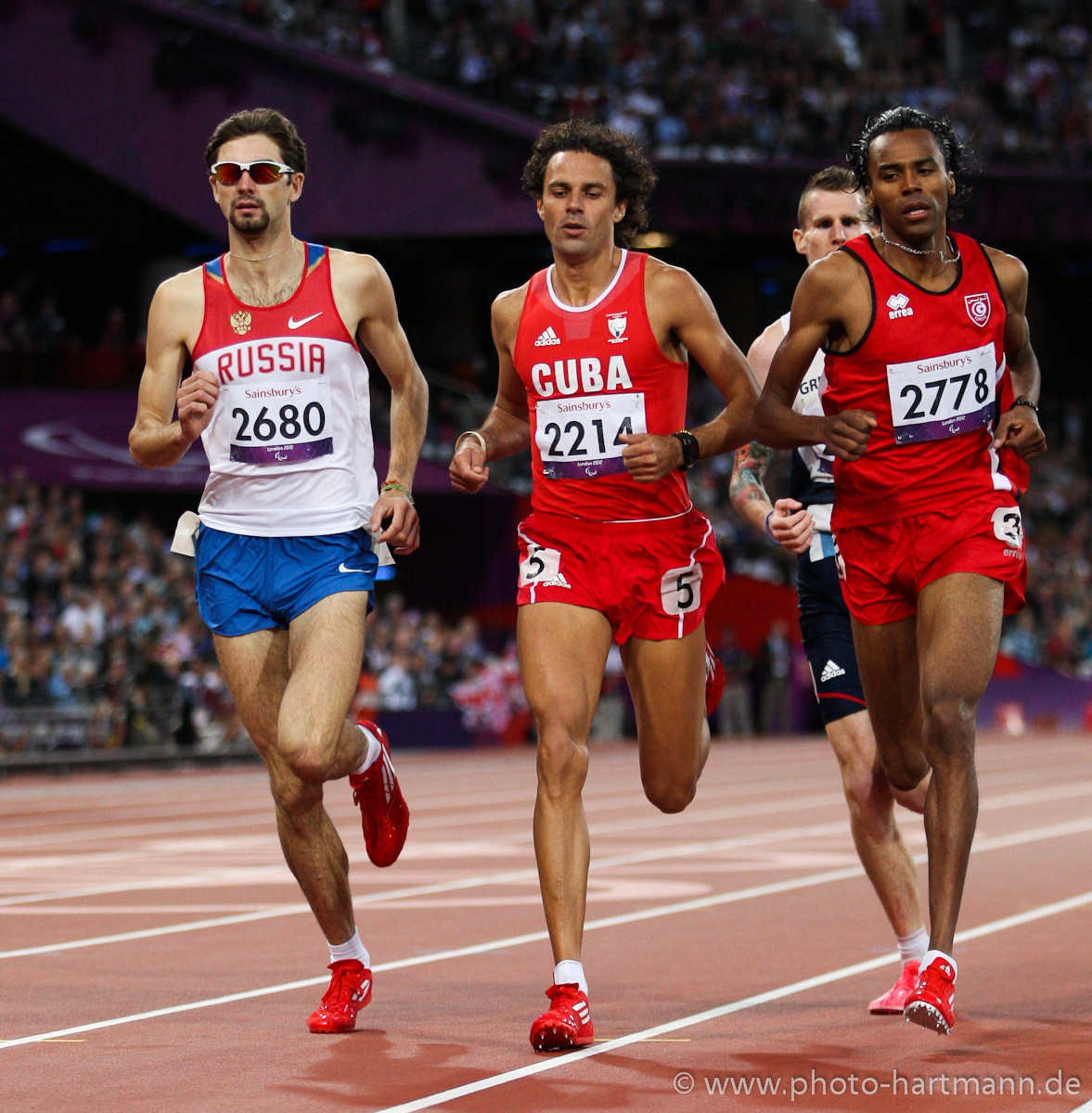 Four runners dressed in shorts and vest competing on a red athletics track.