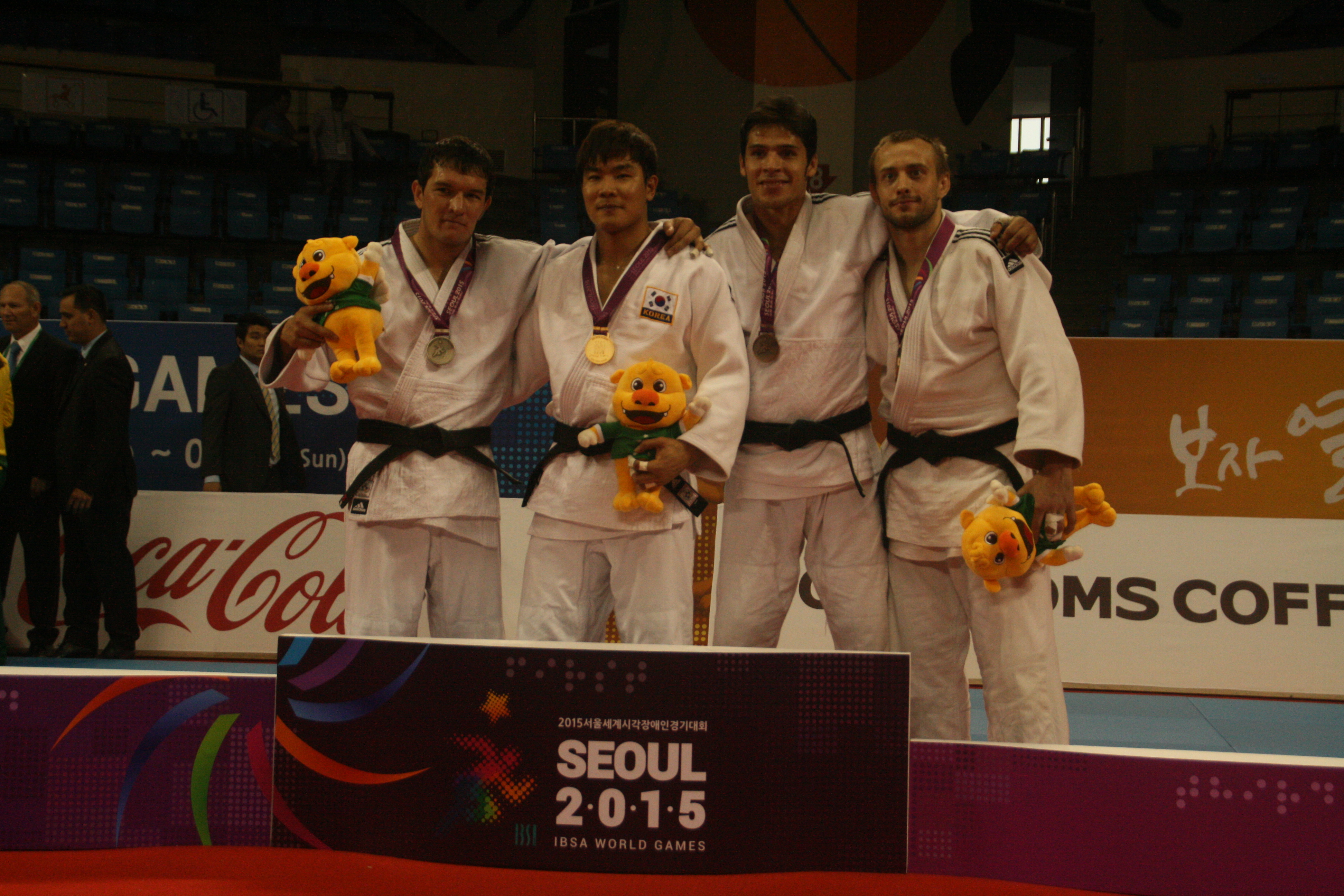 Three men in white training suits stand on a podium with medals