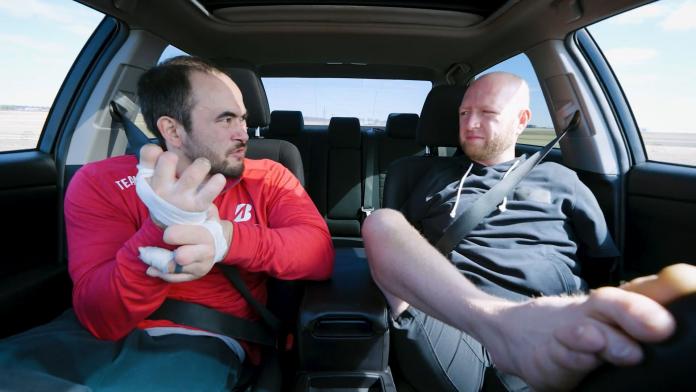 Two Para athletes are sitting in a car. One athlete is driving the car with his foot