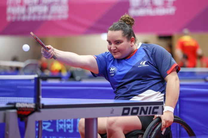 A female Para table tennis player is in action. She is holding the table tennis paddle in her right hand while holding on to the tires of her wheelchair with her left hand.