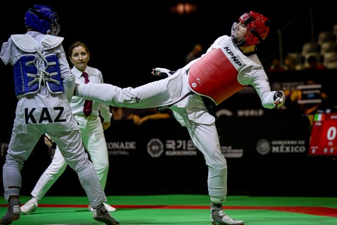 Two male Para taekwondo athletes fight in a match. An athlete wearing a red head gear kicks his opponent wearing a blue head gear.