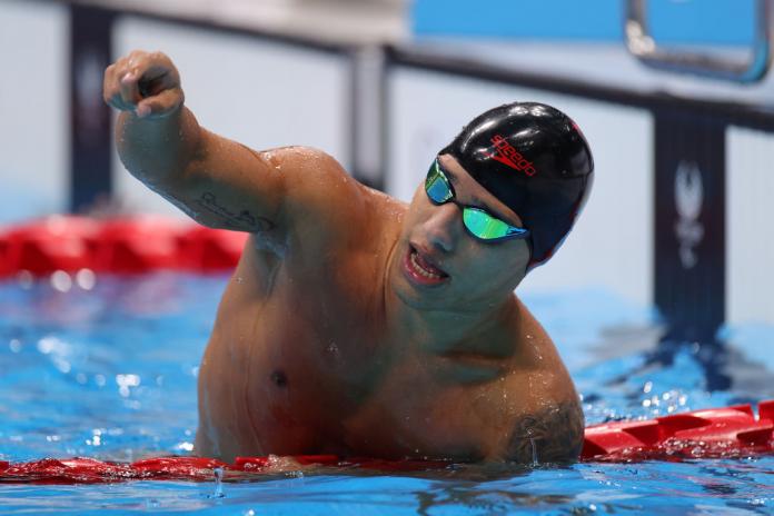 A male Para swimmer raises his right hand in the pool after a competition