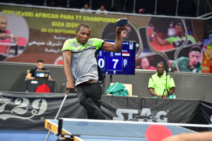 A male Para table tennis player in action. He is holding a cane with his right hand and a table tennis paddle with his left.