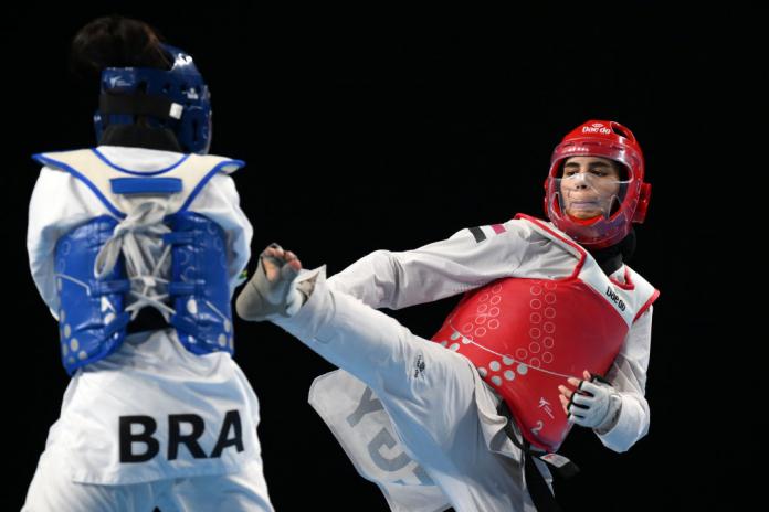 Two female Para taekwondo athletes fight in a match. The athlete wearing a red head gear kicks her opponent wearing a blue head gear.