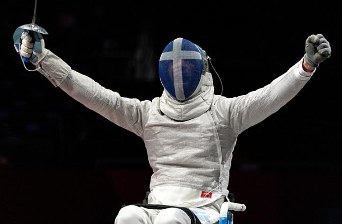 A wheelchair fencing athlete wearing a mask reacts after a match by raising both arms.