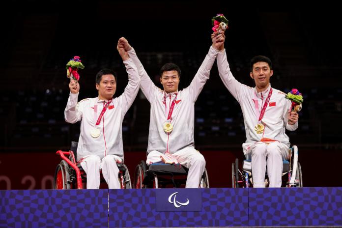 Three Chinese men's wheelchair fencers wearing a white uniform raise their arms after receiving their gold medals