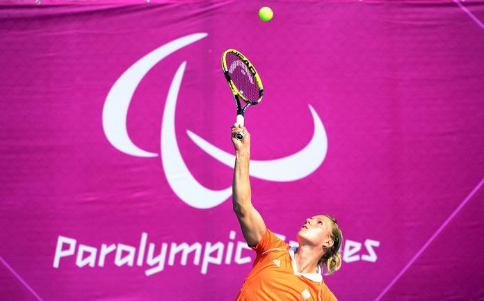 A female wheelchair tennis athlete returns a shot in front of a banner of the Paralympic symbol and the words Paralympic Games