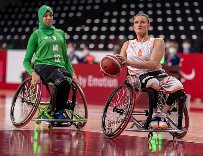 A female wheelchair basketball player carries the ball during a match. A female player from the opposing team is behind her.