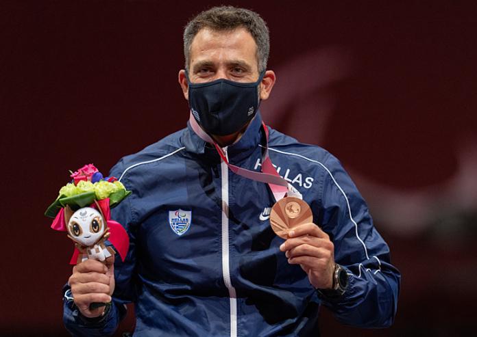 A male wheelchair fencer wearing a face mask poses for a photo after receiving a bronze medal