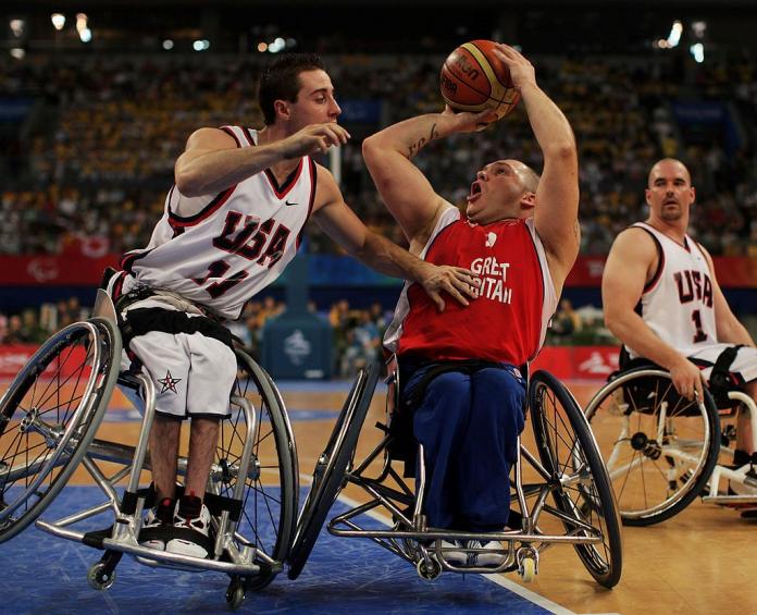 Two male wheelchair basketball players are in action. One player is attempting a throw, while another player is leaning in to block.