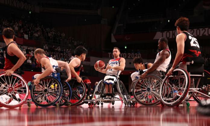 A male wheelchair basketball player is carrying the ball while surrounded by other players