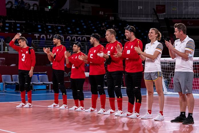 Six men's goalball athletes and two staff stand in a line on court and clap their hands