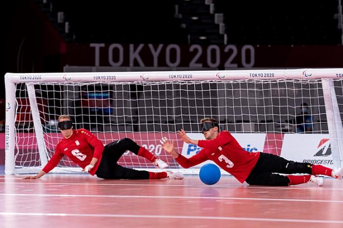 Two male goalball athletes are blocking the ball in front of a net