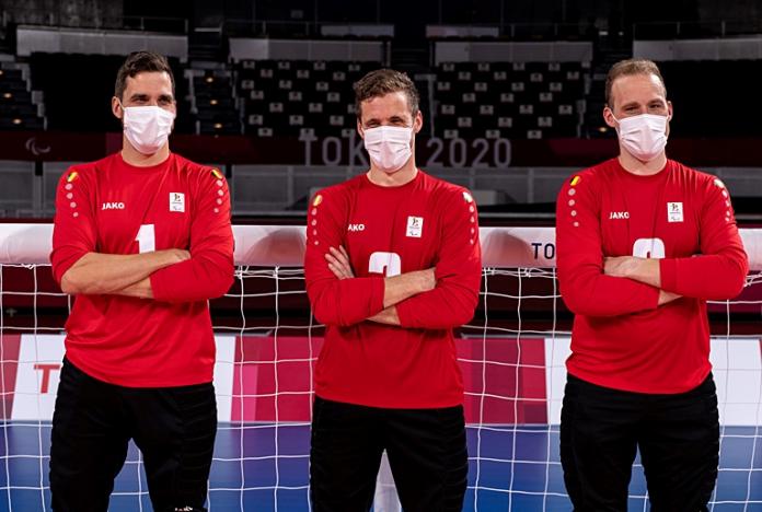 Three goalball athletes wearing red uniform and face masks cross their arms in front of a goalball net