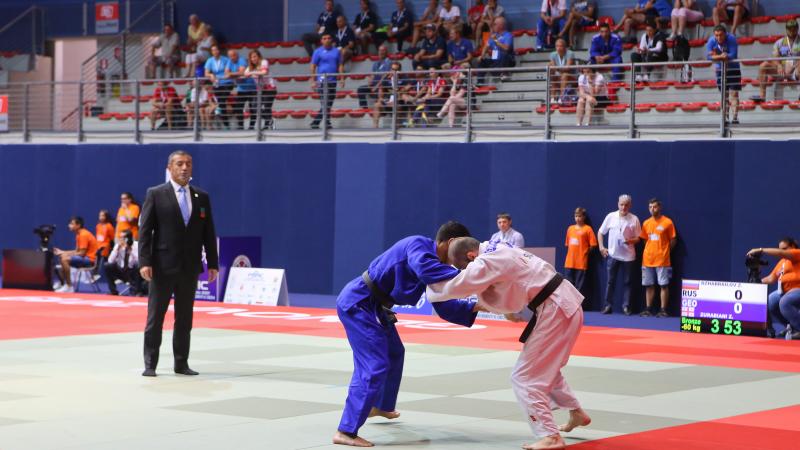 Russian and Georgian judokas grab each other on the tatami as the judge and spectators watch