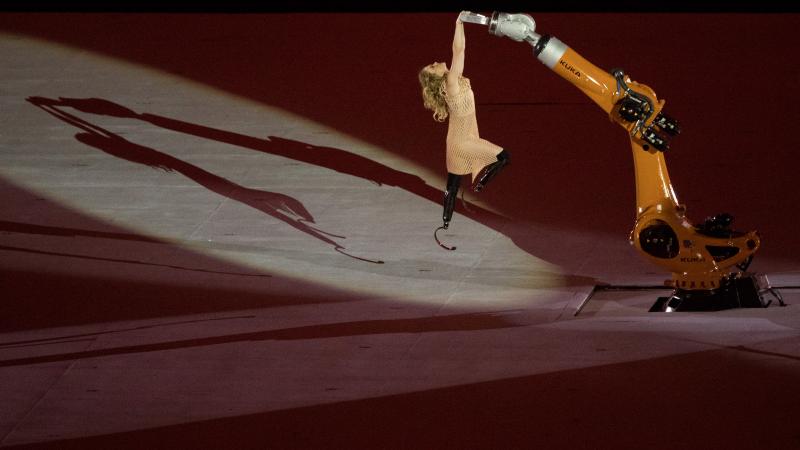 a female with prosthetic legs being raised into the air holding onto a robot arm