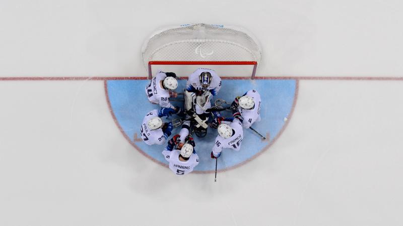 A group of six Para ice hockey players huddle together in front of the goal