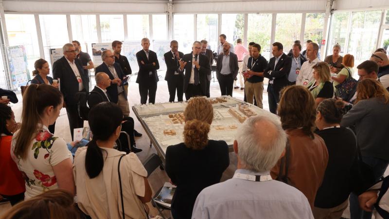 Officials look at a miniature model of a city landscape while one man speaks in a microphone.