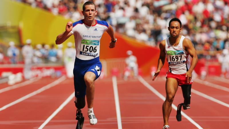 Two men with prosthetic legs sprint on an athletics track at a packed stadium.