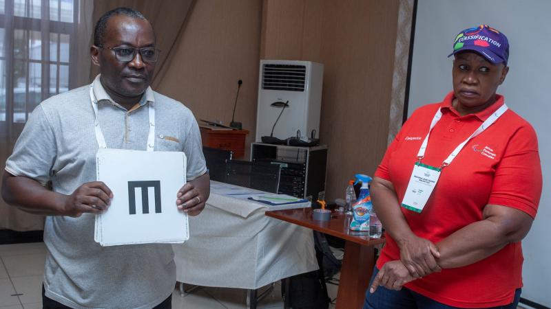 A man holds up charts for testing eyesight while a female supervisor looks on.