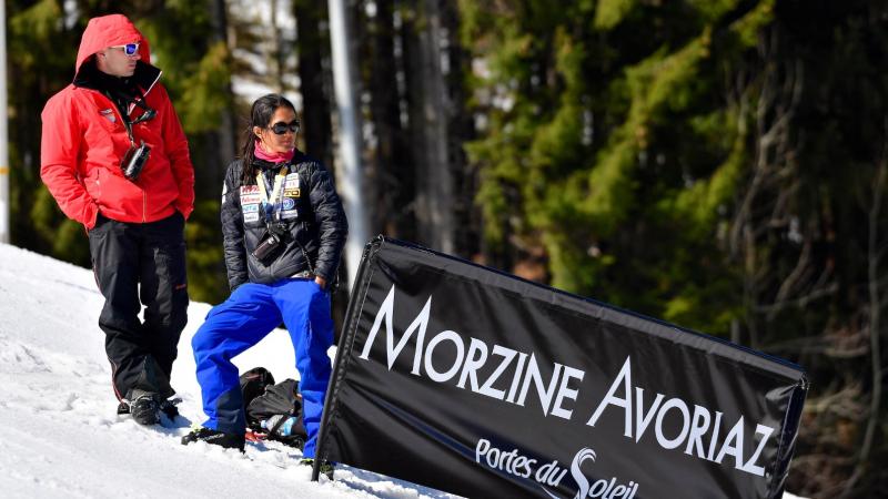 A female coach stands on the snow, next to a male staff and a sign that reads "Morzine Avoriaz"