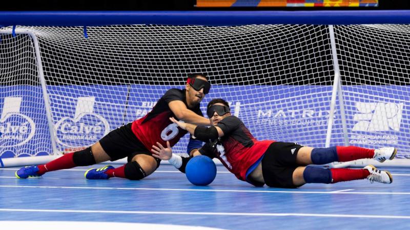 Two male athletes competing in goalball. They are blocking a blue ball in front of the net