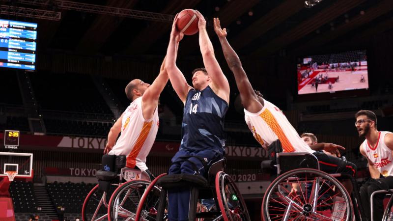A male wheelchair basketball player wearing a blue jersey tries to shoot, while two players wearing a white jersey are reaching out to block him.