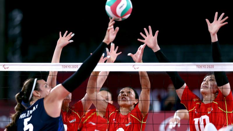 A female athlete tips the ball over the net, while three players try to block it
