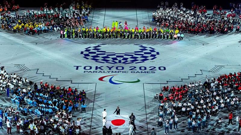 About five people carry the Japanese flag into a stadium during the Closing Ceremony of the Tokyo 2020 Paralympics. The Tokyo 2020 emblem and the Paralympic symbol is depicted on the floor.