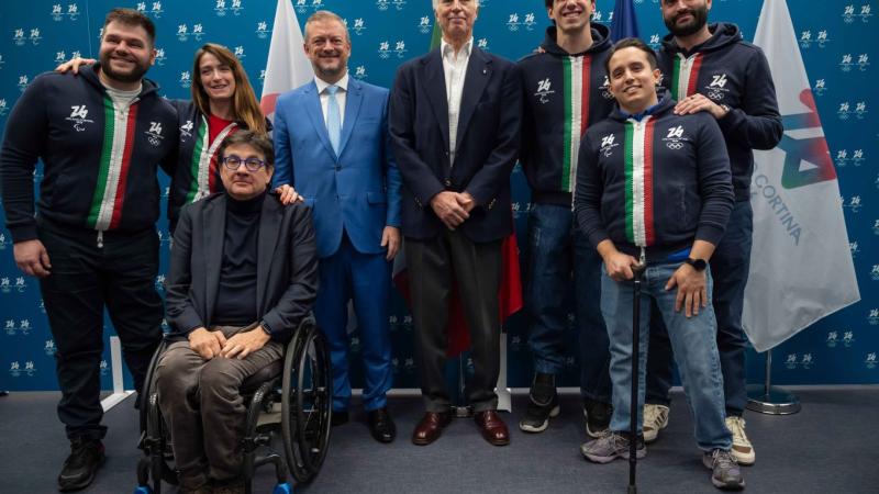 Eight people, including Paralympic athletes and officials, pose for a photograph in front of a blue banner with logos of the Milano Cortina 2026 Games