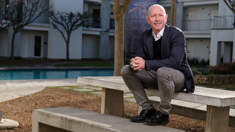 Wayne Derman sits on a stone bench and smiles at the camera