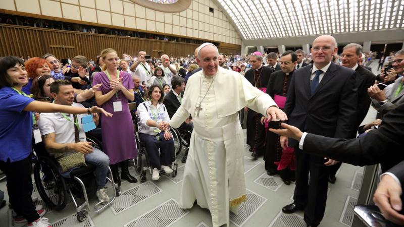 Man in white robe walks through a crowd of people, some of them in wheelchairs