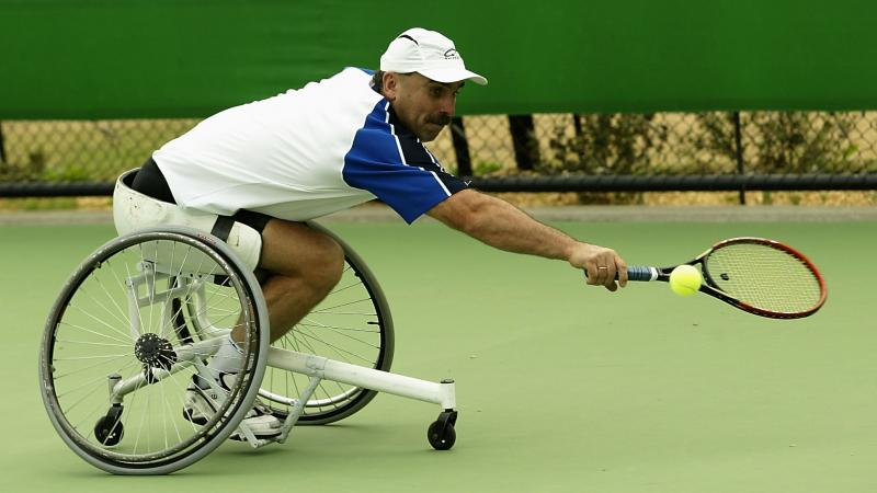 Man in wheelchair reaches to try and hit a tennis ball with his racquet 