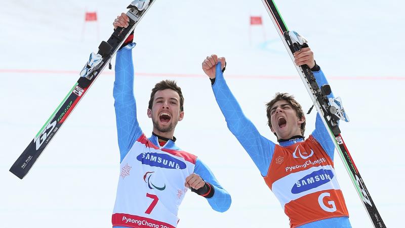 a male vision impaired skier and his guide celebrate by holding up their skis