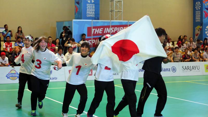 Japanese female goalball players run across the court holding a Japan flag