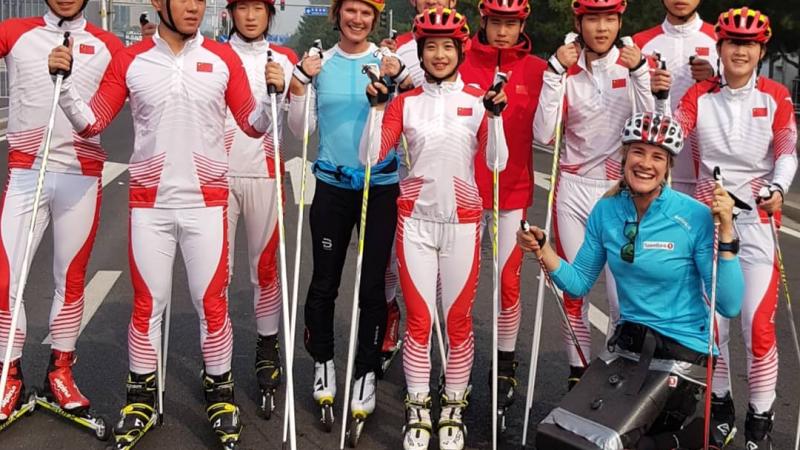 A woman in a sitting rollerski together with a group of ten rollerskiers