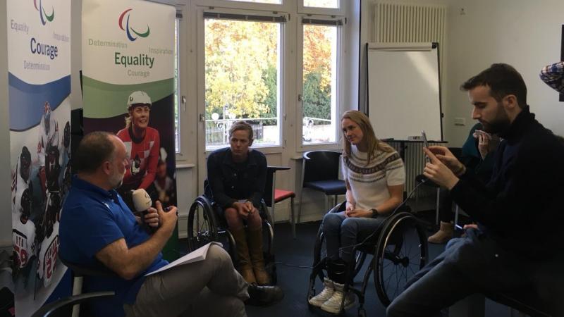 two athletes in wheelchairs watching as a man with a microphone is filmed on a phone